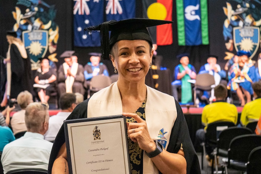 A woman wearing a university cap and gown stands smiling, holding her certificate.
