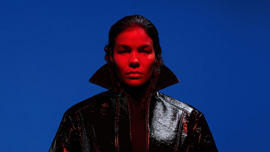 Image of woman with red light on her face and wearing a coat with a popped collar over a blue  background