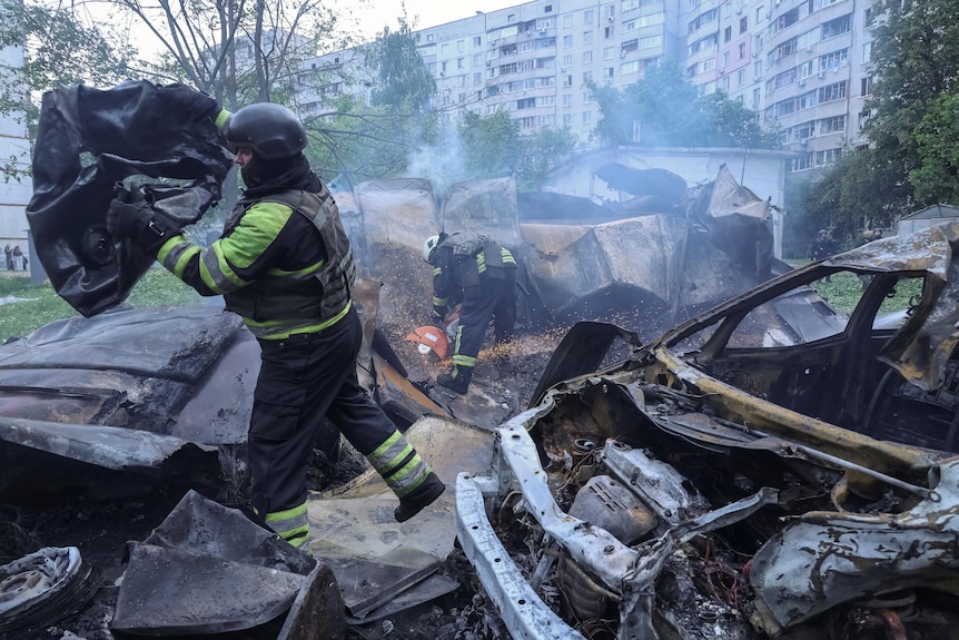 Two men in black and yellow protective gear sift through the smoking wreckage of what look like cars in a street.