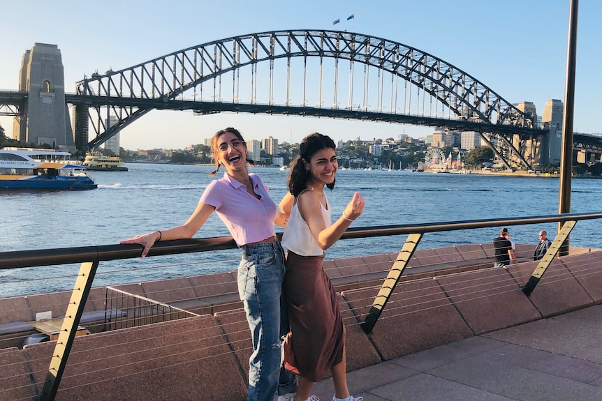 Two women smile for the camera while standing by a railing in front of the Sydney Harbour Bridge.