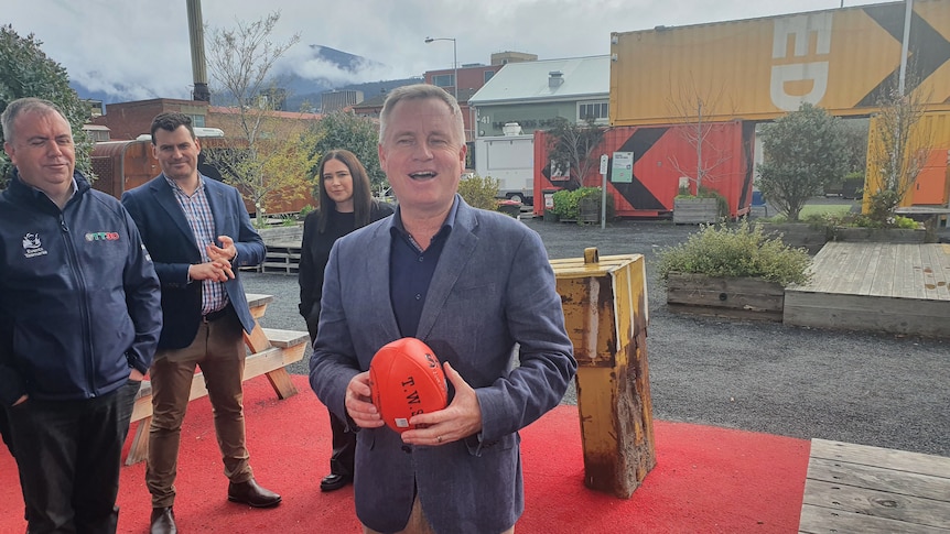 A middle-aged man in grey jacket smiles as he holds orange AFL ball in front of buildings