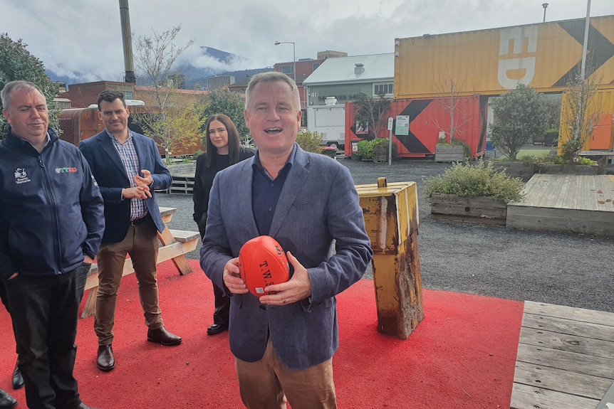 A middle-aged man in grey jacket smiles as he holds orange AFL ball in front of buildings