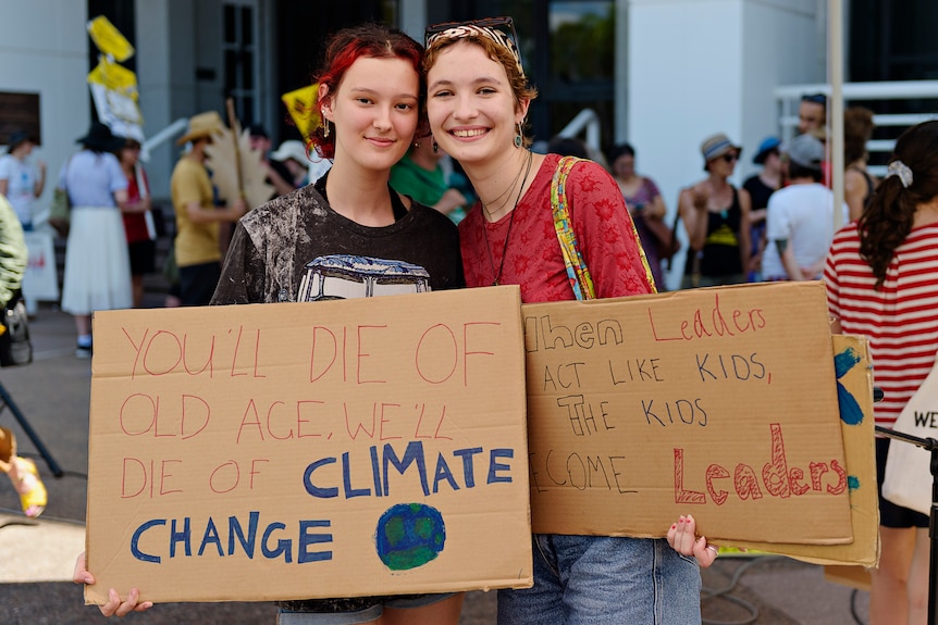 Two young women stand together smiling at the camera holding carboard signs with climate slogans, in front of a crowd. 