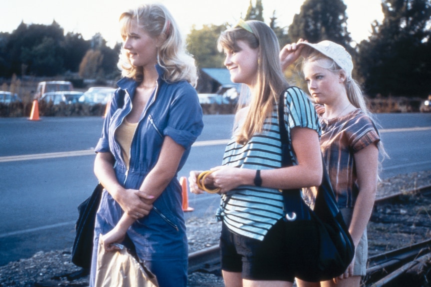 Film still showing three young blonde girls standing in a line by the side of the road, first two smiling.