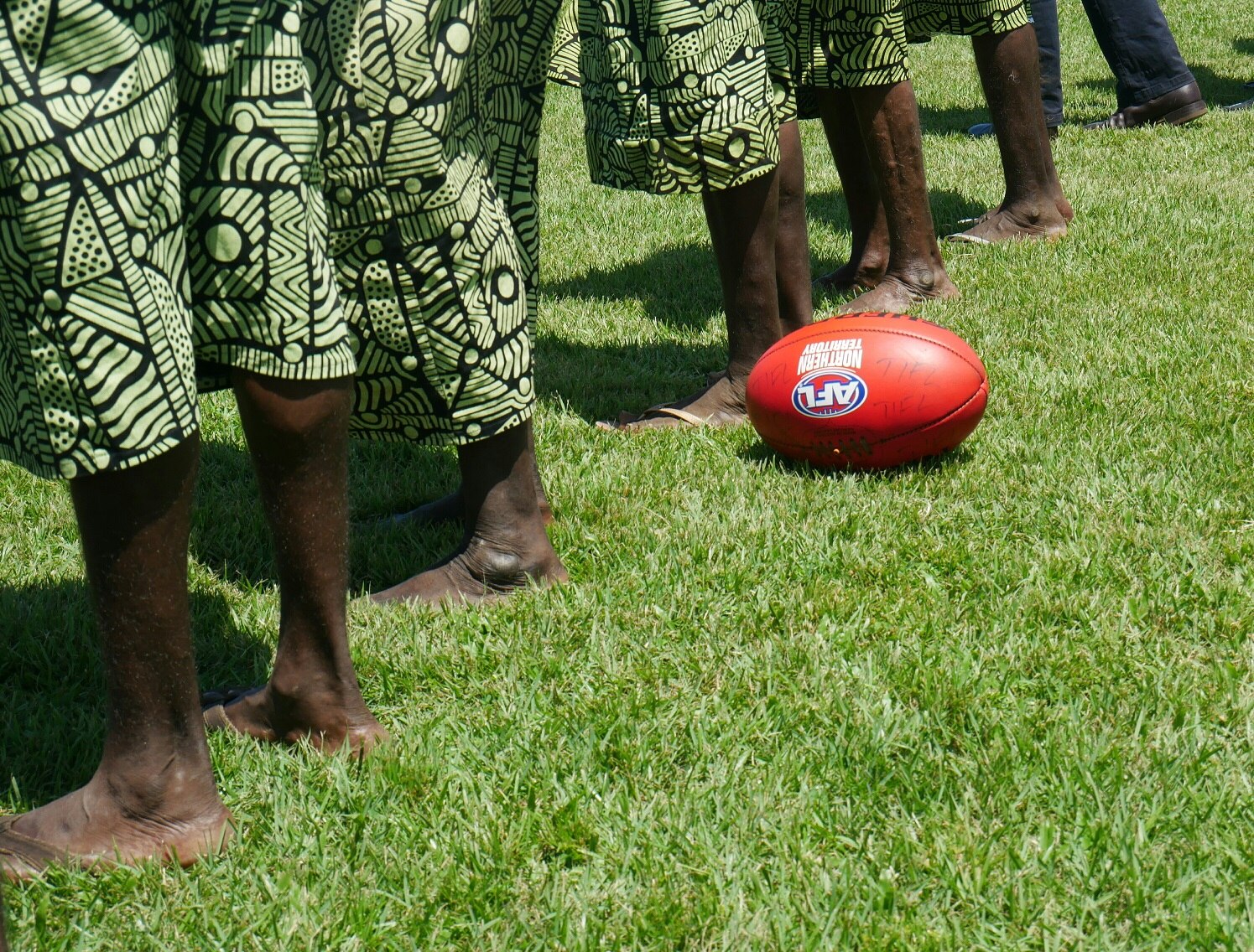 Indigenous men and women stand on a football field next to an AFL ball.