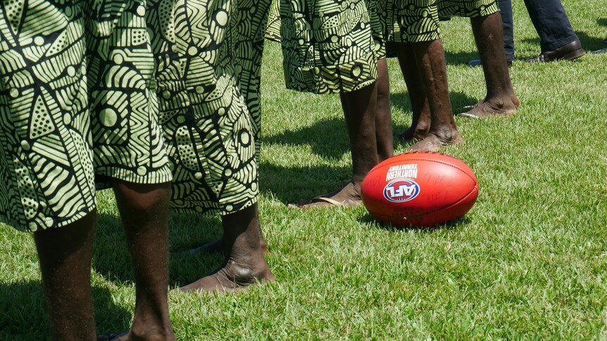 Indigenous men and women stand on a football field next to an AFL ball.