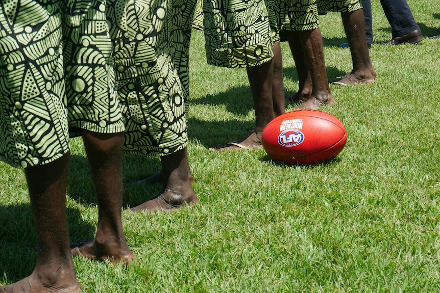 Indigenous men and women stand on a football field next to an AFL ball.