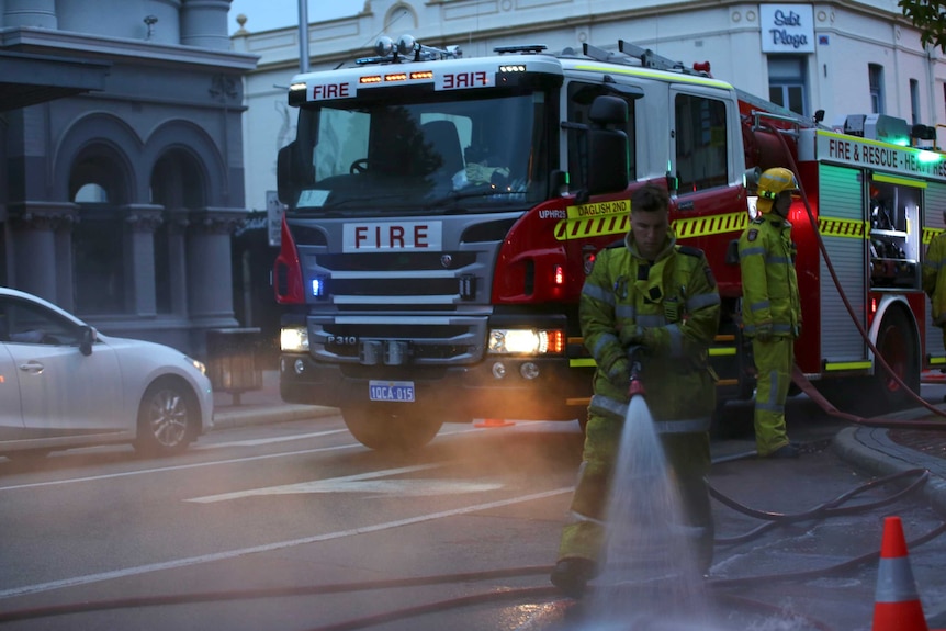 A fire fighter with a hose and and another fire fighter standing by a fire truck in a street