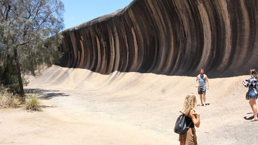 Three tourists stand next to Wave Rock on a clear sunny day.