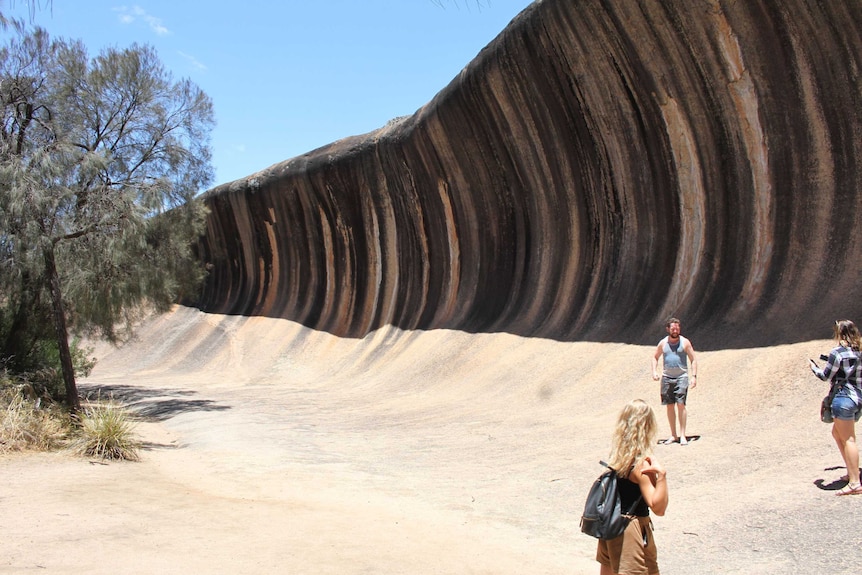 Three tourists stand next to Wave Rock on a clear sunny day.