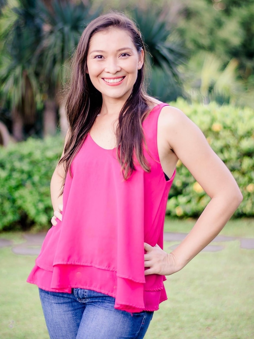 A woman with long brown hair smiles wearing a bright pink top and denim jeans.