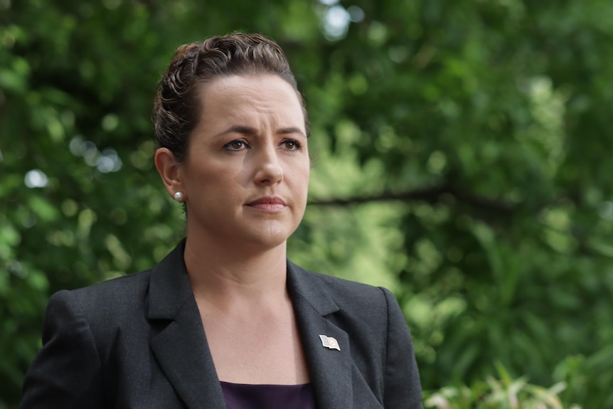 A woman standing in front of a microphone and looking serious, in front of greenery.