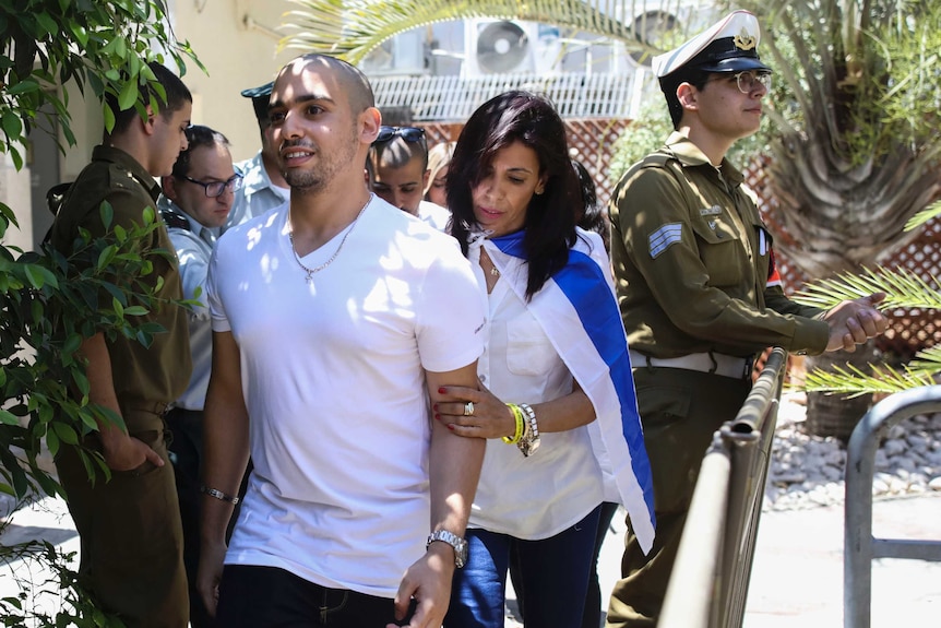 Elor Azaria walks past guards with his mother holding his arm.