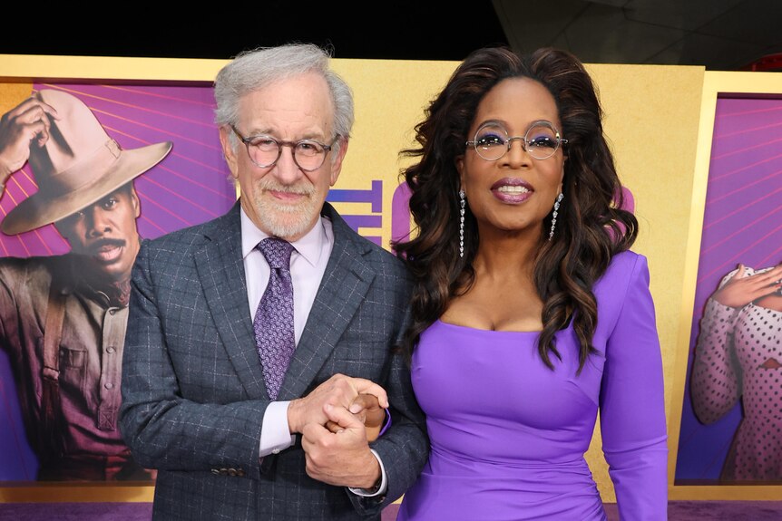 Steven Spielberg in a suit with purple tie and Oprah Winfrey wearing a purple dress and reading glasses