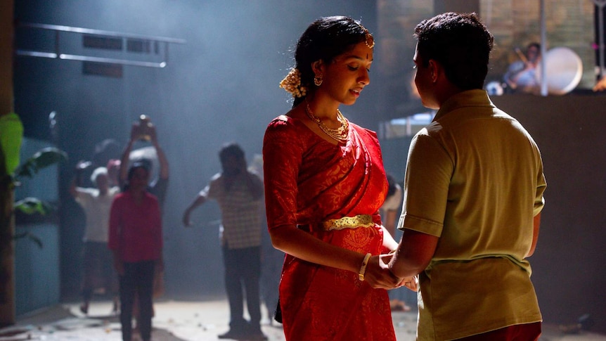 A backlit woman on stage  in a traditional Sri Lankan outfit holds hands with a man facing away from the camera.