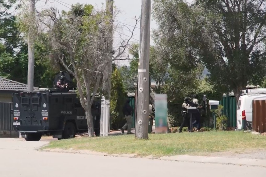 A wide shot showing a WA Police armoured vehicle outside a house on a quiet street with officers walking towards a house.