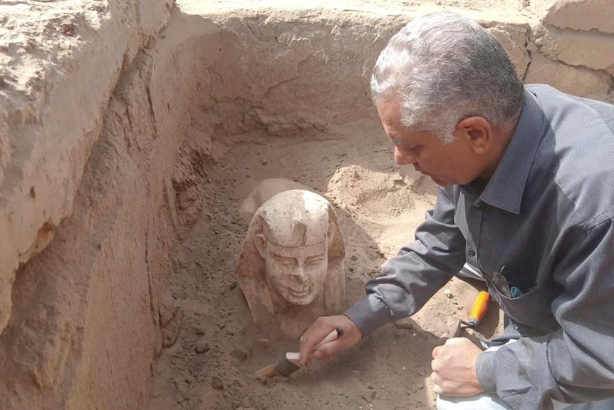 An older Egyptian man in a grey shirt with grey hair brushes the ground near the protruding head of a small Sphinx statue.