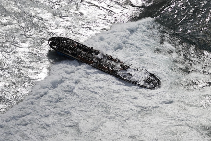 A long tanker type vessel is completely destroyed with fire burning holes in the hull