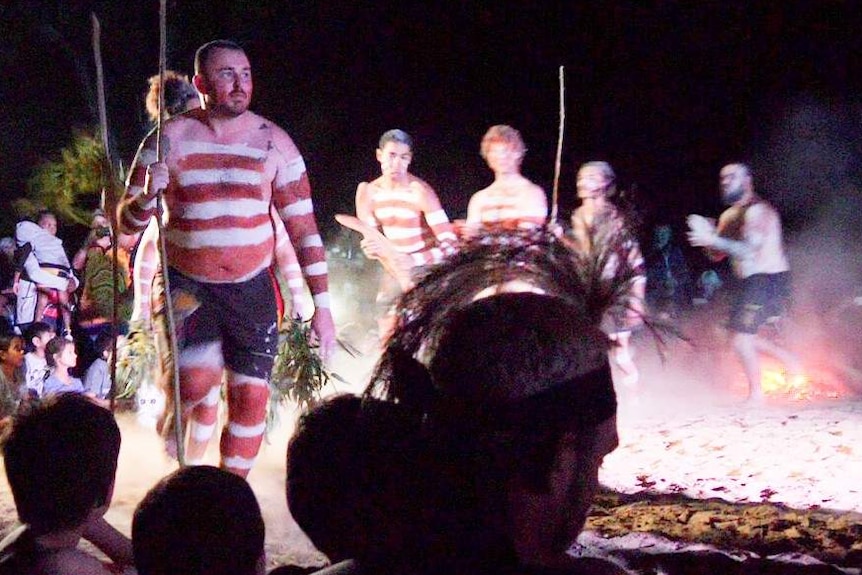 A line of Aboriginal dancers painted in white and ochre move around a dance circle at night.