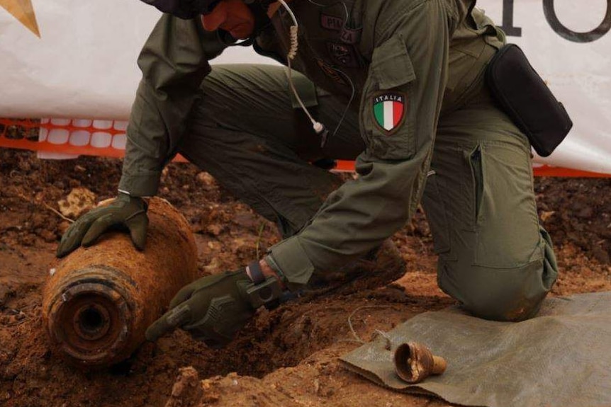 A man in a green military uniform wearing a helmet kneels over a rusty, dirty bomb.