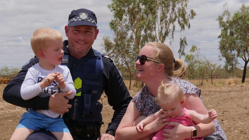 Kynuna police officer in charge Senior Constable Graeme Ferguson kneels in the red dirt with his wife and two young children.