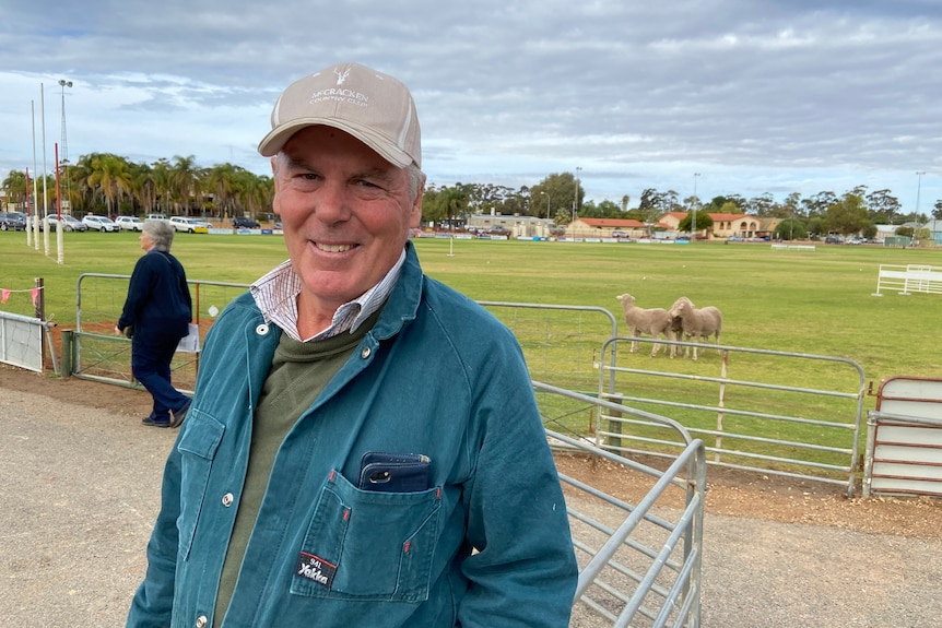 A man in a cap and overalls stands at a pen on an oval in front of some sheep.