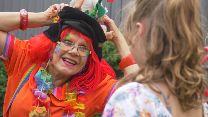 A woman dressed in very bright colourful clothes as a clown, with her hands on her head, making a child smile.