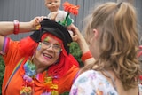 A woman dressed in very bright colourful clothes as a clown, with her hands on her head, making a child smile.
