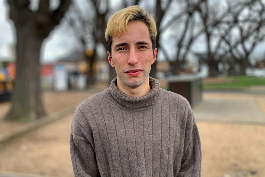 A man in his 20's with bleached hair stands in a park on a winter day. He's wearing a grey jumper