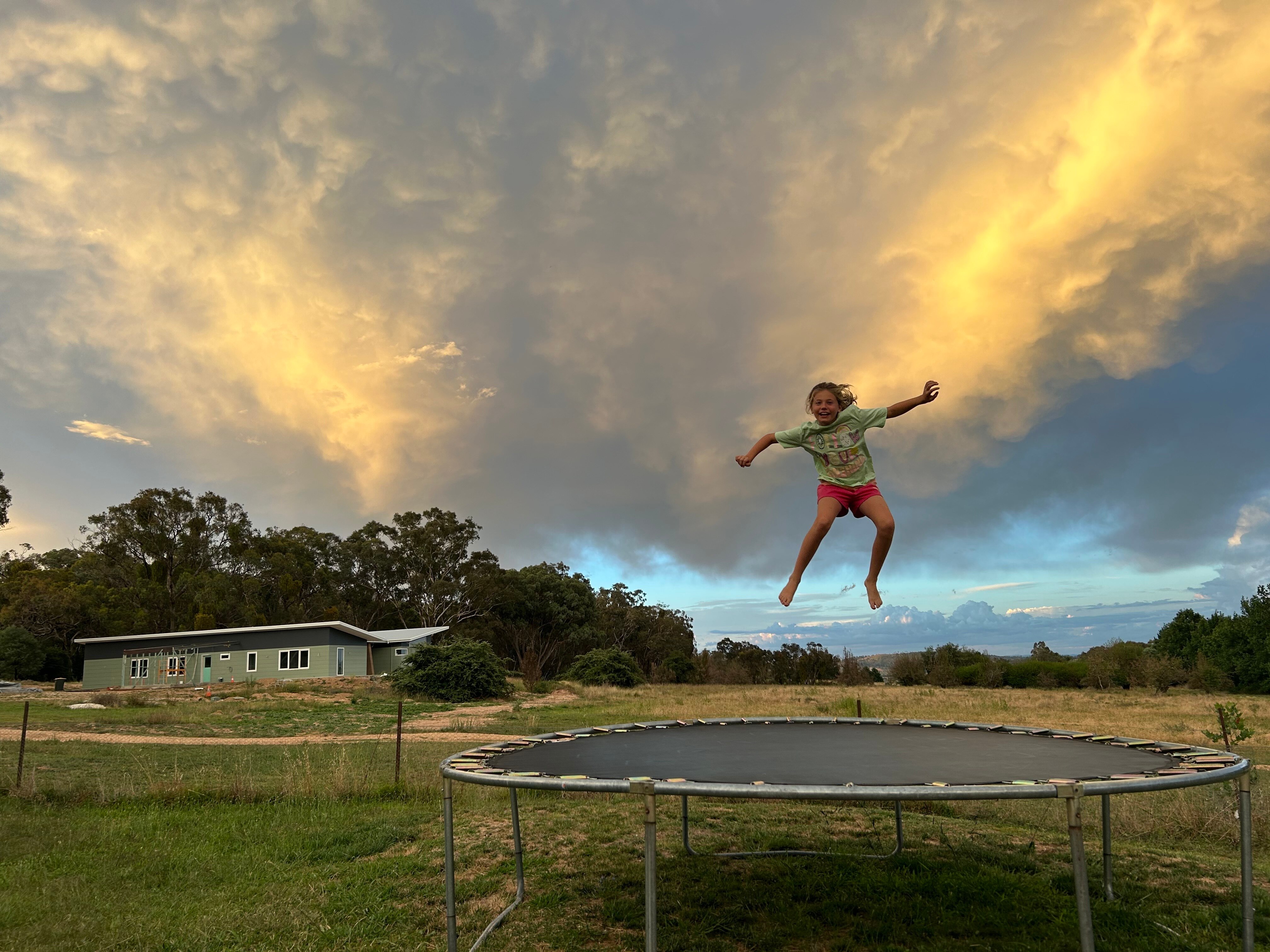 A young girl jumps on a trampoline on a property on an overcast day, a new home being built is behind her in the distance.