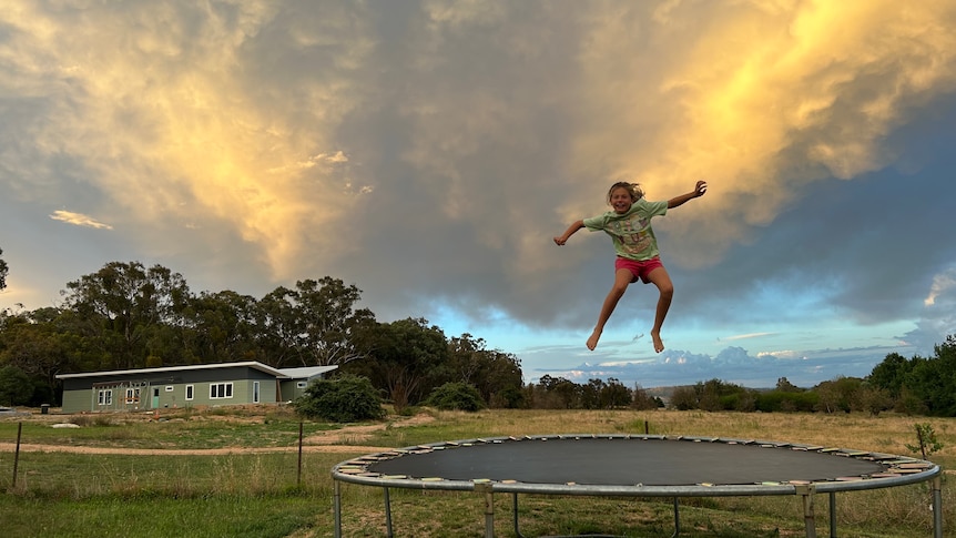 A young girl jumps on a trampoline on a property on an overcast day, a new home being built is behind her in the distance.