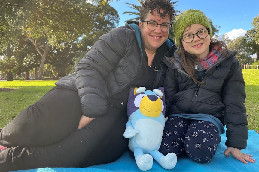 A heavyset middle aged woman with short hair and glasses smiles and leans close to a child wearing a beanie and glasses.