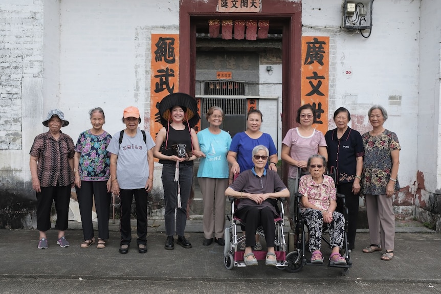 Rainbow Chan, a young Chinese Australian woman stands holding a camera among 10 elderly Weitou women.