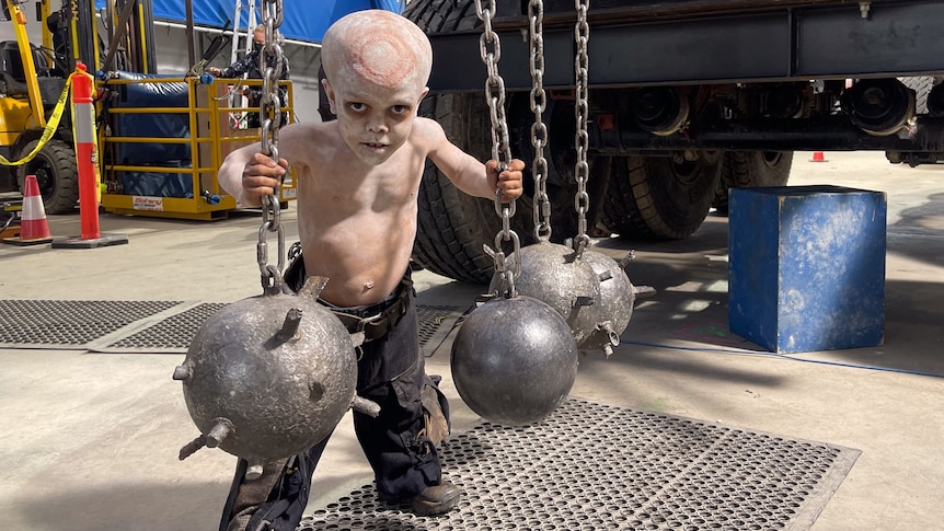 Quaden in Mad Max costume, baggy black pants, no shirt and a red ring design on bald head. He holds hanging chains spiky balls