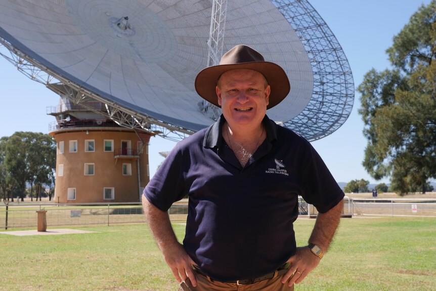 A middle aged man in an akubra standing in front of a observatory dish 