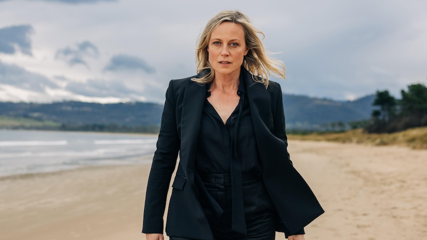 A woman stands on a beach wearing a black suit and black shirt.