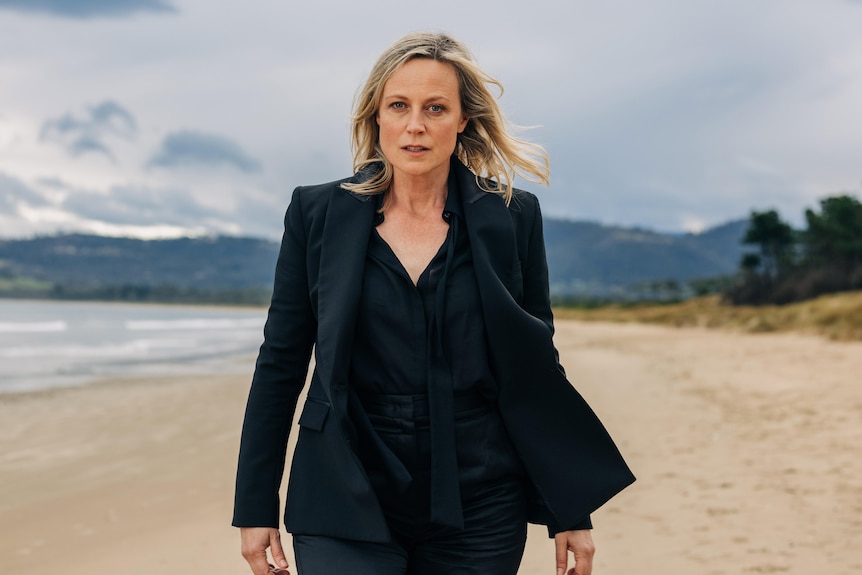 A woman stands on a beach wearing a black suit and black shirt.