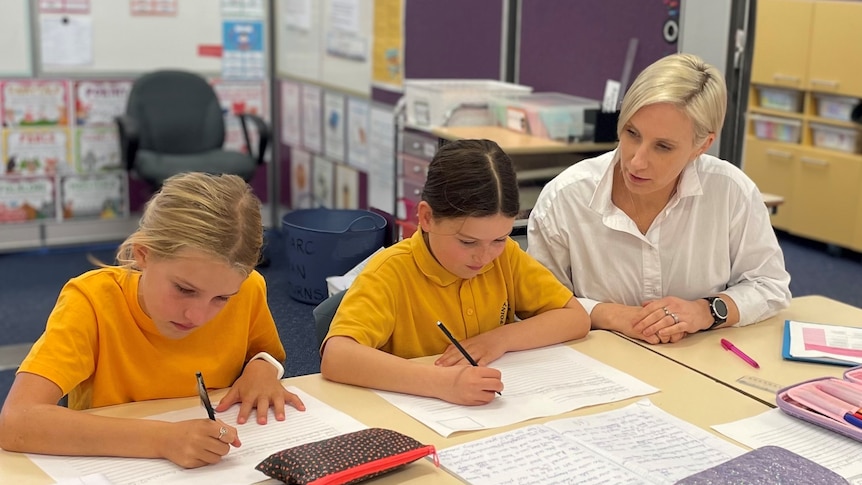 A teacher sits at a table helping two primary school aged students with their work.