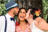 A just-married bride and groom each kiss the celebrant on a cheek at an outdoor ceremony