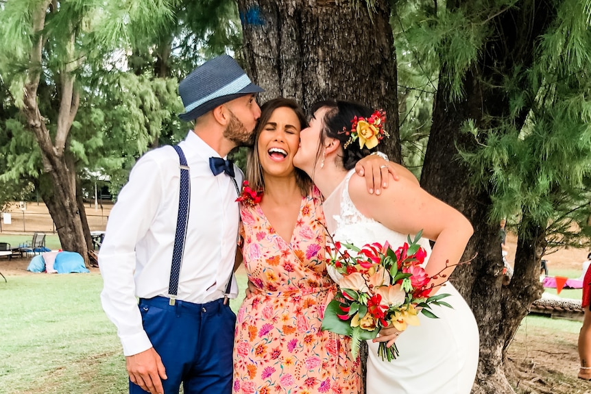 A just-married bride and groom each kiss the celebrant on a cheek at an outdoor ceremony