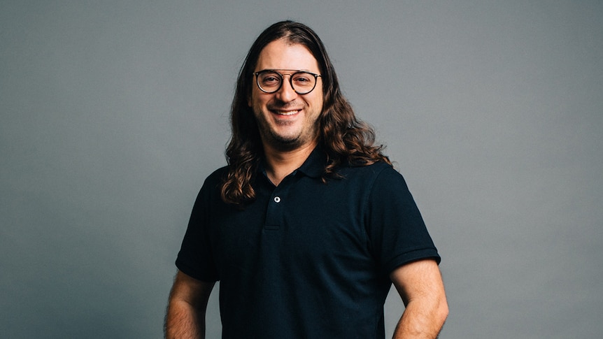 Matt Gudinski with long hair and glasses wearing all black, smiling at the camera while standing with his hands in his pockets