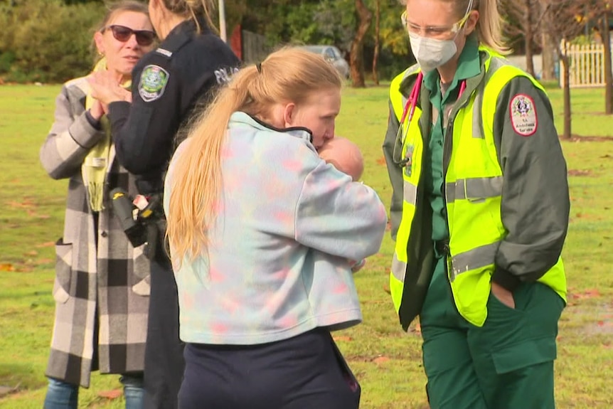 Une femme portant un pull gris embrasse un bébé serré dans ses bras alors qu'un ambulancier sourit et regarde