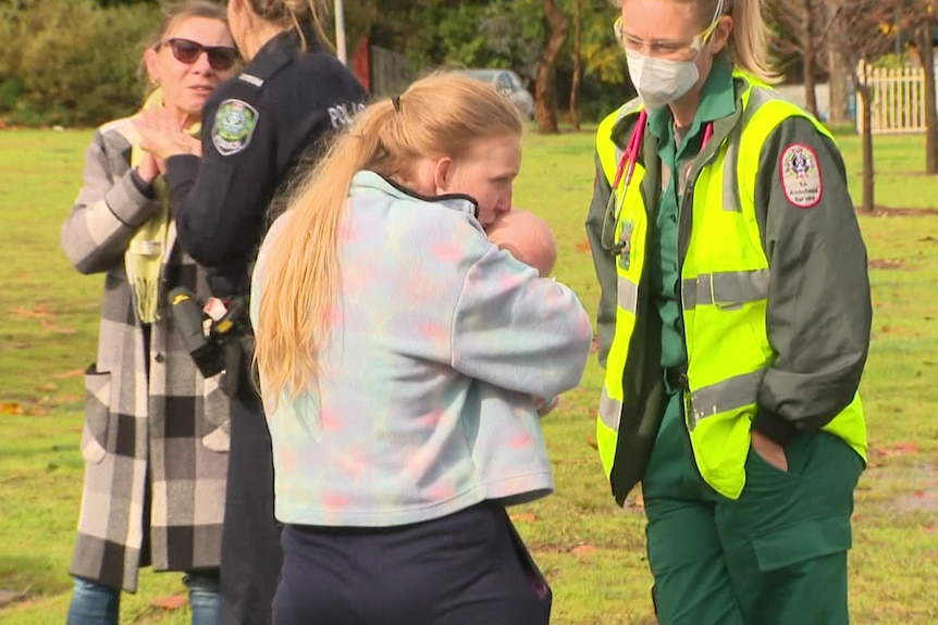 A woman in a grey jumper kisses a baby held tight in her arms as a paramedic smiles and looks on