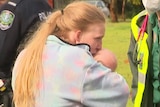 A woman in a grey jumper kisses a baby held tight in her arms as a paramedic smiles and looks on