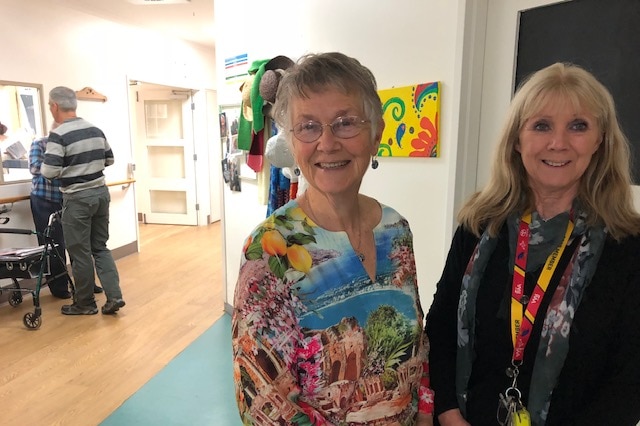 Sally Goode and Sandy Eisner at a corridor at an aged care facility, with two men with walker in background.