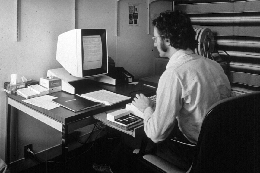 A black and white photo of a man with long sideburns sitting at a small, old-timey computer.