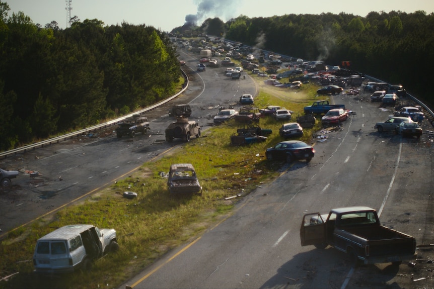 More than 30 abandoned cars and trucks litter a highway with no humans in sight. Some cars are smoking.
