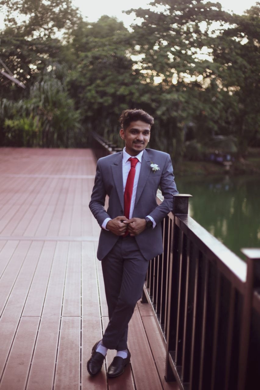 A young man in a suit leans against a bridge over a body of water. 