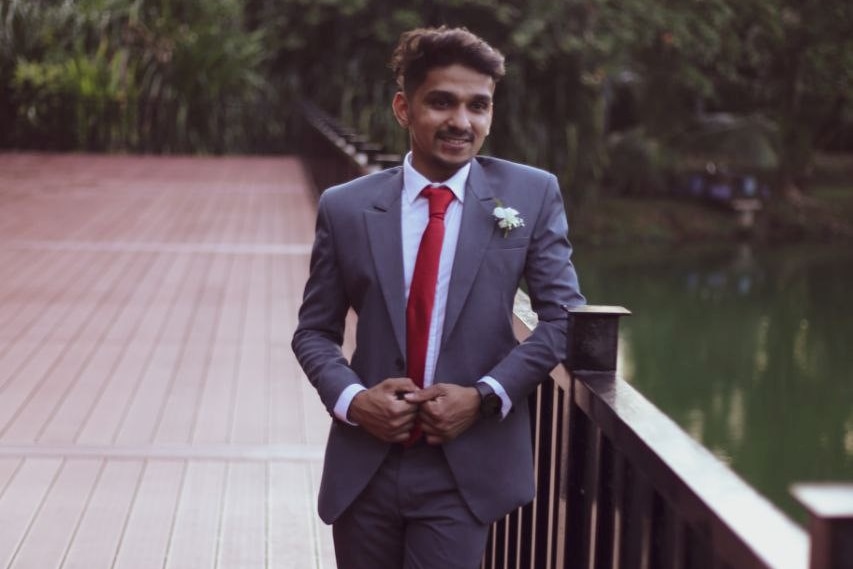 A young man in a suit leans against a bridge over a body of water. 