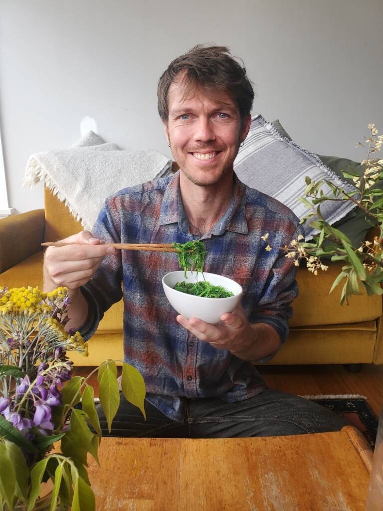 Man splies at camera holding chopsticks and a bowl of seaweed. 